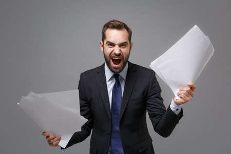 Angry irritated young business man in classic black suit shirt tie posing isolated on grey background. Achievement career wealth business concept. Mock up copy space. Holding work documents screaming.の写真素材