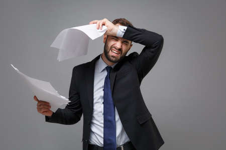 Exhausted young business man in classic black suit shirt tie posing isolated on grey background. Achievement career wealth business concept. Mock up copy space. Hold work documents put hand on head.の写真素材