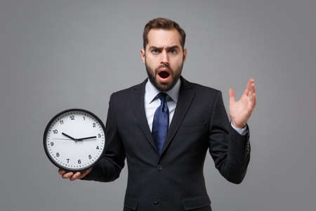 Shocked young bearded business man in classic black suit shirt tie posing isolated on grey background. Achievement career wealth business concept. Mock up copy space. Holding clock, spreading hands.の写真素材