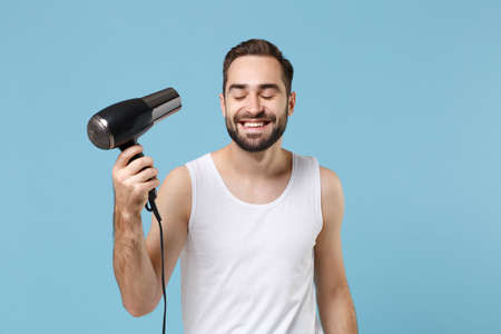 Bearded fun young man 20s years old in white shirt hold blow dry hair isolated on blue pastel wall background, studio portrait. Skin care healthcare cosmetic procedures concept. Mock up copy space.の写真素材