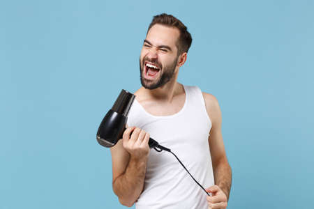 Bearded fun young man 20s years old in white shirt hold blow dry hair isolated on blue pastel wall background, studio portrait. Skin care healthcare cosmetic procedures concept. Mock up copy space.の写真素材