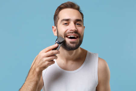 Close up bearded young man 20s years old in white shirt hold electric razor isolated on blue pastel background, studio portrait. Skin care healthcare cosmetic procedures concept. Mock up copy space.の写真素材