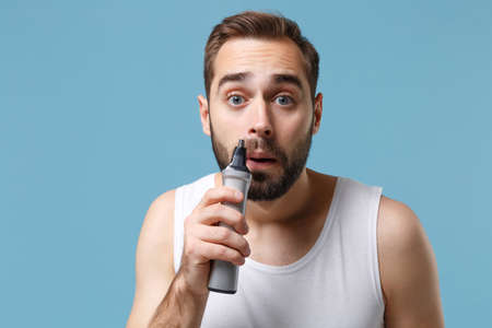 Close up bearded young man 20s years old in white shirt hold electric trimmer isolated on blue...