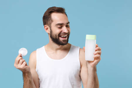 Bearded young man 20s years old in white shirt hold in hand cotton pad isolated on blue pastel wall background, studio portrait. Skin care healthcare cosmetic procedures concept. Mock up copy space.の写真素材