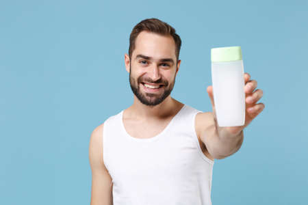 Bearded young man 20s years old in white shirt hold in hand bottle tonic isolated on blue pastel wall background, studio portrait. Skin care healthcare cosmetic procedures concept. Mock up copy space.の写真素材