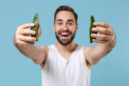 Close up bearded young man 20s years old in white shirt hold half of avocado isolated on blue pastel background, studio portrait. Skin care healthcare cosmetic procedures concept. Mock up copy space.の写真素材