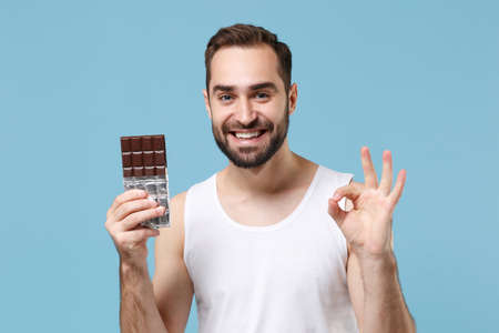 Bearded young man 20s years old in white shirt hold in hand chocolate bar isolated on blue pastel wall background, studio portrait. Skin care healthcare cosmetic procedures concept. Mock up copy spaceの写真素材