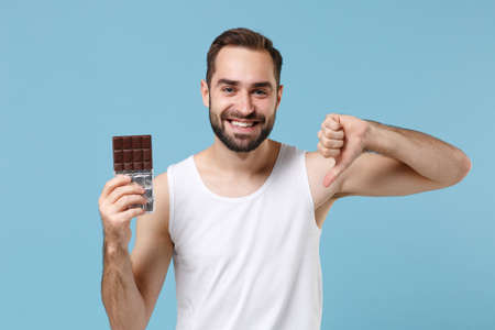 Bearded young man 20s years old in white shirt hold in hand chocolate bar isolated on blue pastel wall background, studio portrait. Skin care healthcare cosmetic procedures concept. Mock up copy spaceの写真素材