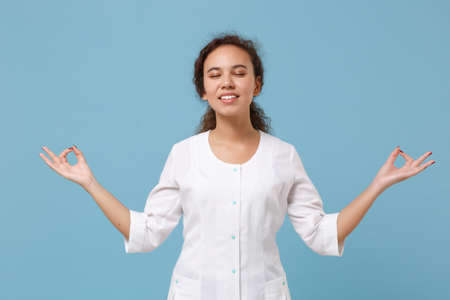 African american doctor woman isolated on blue background. Female doctor in medical gown hold hands in yoga gesture, relax meditating. Healthcare personnel health medicine concept. Mock up copy space.の写真素材