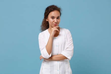Beautiful african american doctor woman isolated on blue background. Female doctor in white medical gown put hand prop up on chin. Healthcare personnel health medicine concept. Mock up copy space.の写真素材