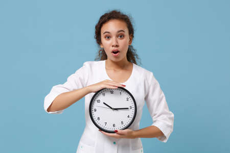 Shocked african american female doctor woman in white medical gown holding round clock isolated on blue background studio portrait. Healthcare personnel medicine health concept. Mock up copy space.の写真素材