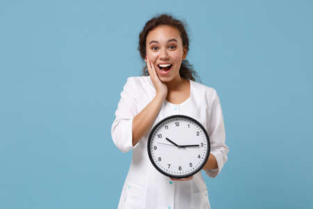 Excited african american female doctor woman in white medical gown holding round clock isolated on blue background studio portrait. Healthcare personnel medicine health concept. Mock up copy space.の写真素材