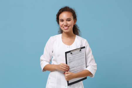 Smiling african american female doctor woman in white medical gown hold clipboard with medical documents isolated on blue background. Healthcare personnel medicine health concept. Mock up copy space.の写真素材