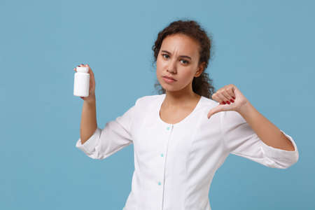 African american female doctor woman in medical gown showing thumb down holding tablets, aspirin pills in bottle isolated on blue background. Healthcare personnel medicine concept. Mock up copy space.の写真素材