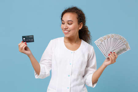 Pretty african american doctor woman isolated on blue background. Female doctor in medical gown hold fan of cash dollar money, credit card. Healthcare personnel medicine concept. Mock up copy space.の写真素材