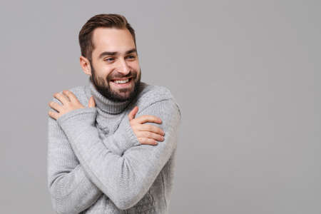 Smiling frozen young man in gray sweater posing isolated on grey background, studio portrait. Healthy fashion lifestyle, cold season concept. Mock up copy space. Holding hands folded, huging himself.の写真素材