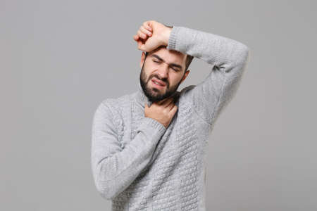 Young bearded man in gray sweater posing isolated on grey background, studio portrait. Healthy lifestyle, ill sick disease treatment, cold season concept. Mock up copy space. Put hand on head, throat.の写真素材
