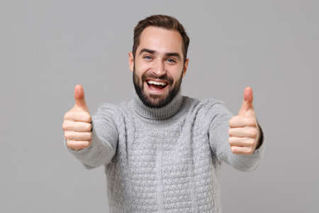 Funny young bearded man in gray sweater posing isolated on grey background, studio portrait. Healthy fashion lifestyle, people emotions, cold season concept. Mock up copy space. Showing thumbs up.の写真素材