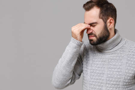 Young man in gray sweater posing isolated on grey background in studio. Healthy lifestyle, ill sick disease treatment, cold season concept. Mock up copy space. Put hand on nose keeping eyes closed.の写真素材