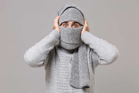 Frozen young man in gray sweater, scarf posing isolated on grey background studio portrait. Healthy fashion lifestyle cold season concept. Mock up copy space. Wrapping in scarf covering face and head.の写真素材