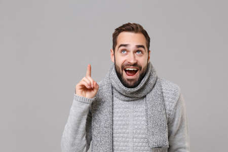 Pensive young man in gray sweater scarf posing isolated on grey background, studio portrait. Healthy fashion lifestyle cold season concept. Mock up copy space. Holding index finger up with great idea.の写真素材