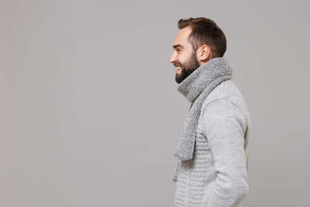 Side view of smiling young man in gray sweater, scarf posing isolated on grey background, studio portrait. Healthy fashion lifestyle, people sincere emotions, cold season concept. Mock up copy space.の写真素材
