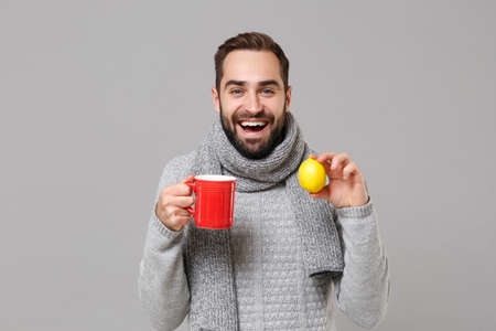 Cheerful young man in gray sweater, scarf posing isolated on grey wall background, studio portrait. Healthy fashion lifestyle, cold season concept. Mock up copy space. Holding red cup of tea, lemon.の写真素材