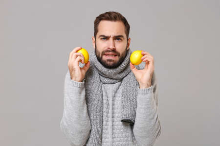 Frozen young man in gray sweater, scarf posing isolated on grey wall background, studio portrait....