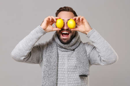 Young man in gray sweater, scarf posing isolated on grey wall background studio portrait. Healthy fashion lifestyle, people emotions, cold season concept. Mock up copy space. Covering eyes with lemon.の写真素材