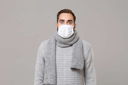 Shocked young man in gray sweater, scarf isolated on grey background studio portrait. Healthy lifestyle, ill sick disease treatment, cold season concept. Mock up copy space. Wearing sterile face mask.の写真素材
