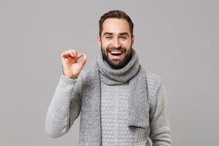 Cheerful young man in gray sweater, scarf isolated on grey wall background. Healthy lifestyle ill sick disease treatment, cold season concept. Mock up copy space. Hold medication tablet, aspirin pill.の写真素材