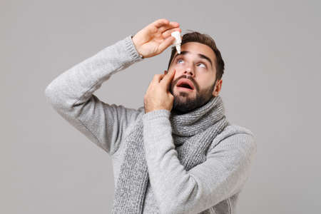 Young man in gray sweater, scarf isolated on grey wall background studio portrait. Healthy lifestyle, ill sick disease treatment, cold season concept. Mock up copy space. Holding, dripping eye drops.の写真素材