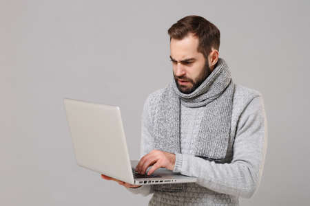 Puzzled young man in gray sweater, scarf isolated on grey background. Healthy lifestyle ill sick disease, online treatment consulting, cold season concept. Mock up copy space. Hold laptop pc computer.の写真素材