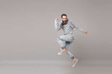 Young bearded man in gray sweater, scarf posing isolated on grey wall background, studio portrait. Healthy fashion lifestyle, cold season concept. Mock up copy space. Jumping, doing winner gesture.の写真素材
