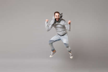 Happy young bearded man in gray sweater, scarf posing isolated on grey background, studio portrait. Healthy fashion lifestyle, cold season concept. Mock up copy space. Jumping, doing winner gesture.の写真素材