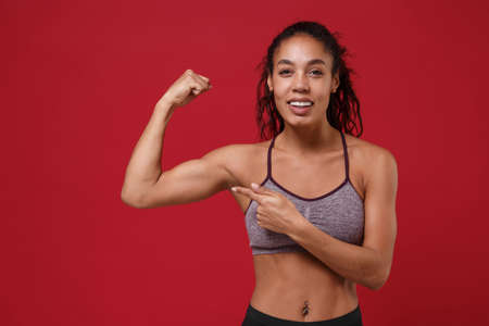 Strong young african american sports fitness woman in sportswear posing working out isolated on red wall background. Sport exercises healthy lifestyle concept. Pointing index finger on biceps muscles.の写真素材