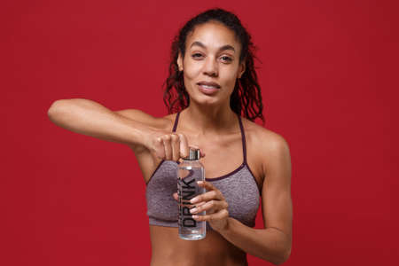 Smiling young african american fitness woman in sportswear posing isolated on red wall background studio portrait. Sport exercises healthy lifestyle concept. Holding water bottle while working out.の写真素材