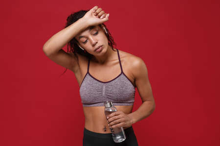 Tired african american fitness woman in sportswear isolated on red background. Sport exercises healthy lifestyle concept. Hold water bottle, working out, listen music with earpods, put hand on head.の写真素材