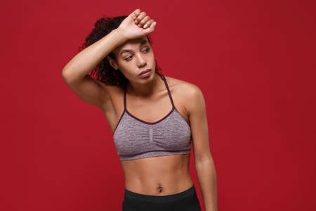 Exhausted young african american sports fitness woman in sportswear posing working out isolated on red wall background. Sport exercises healthy lifestyle concept. Putting hand on head, looking aside.の写真素材