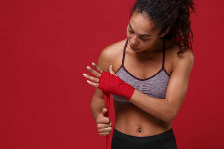 Confident young african american sports fitness woman in sportswear posing working out isolated on red background. Sport exercises healthy lifestyle concept. Reeling up sports bandages on her hands.の写真素材
