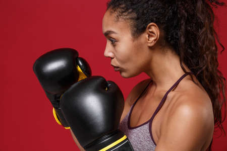 Side view of young african american sports fitness boxer woman in sportswear working out isolated on red background. Sport exercise healthy lifestyle concept. Make boxing exercises in boxing gloves.の写真素材