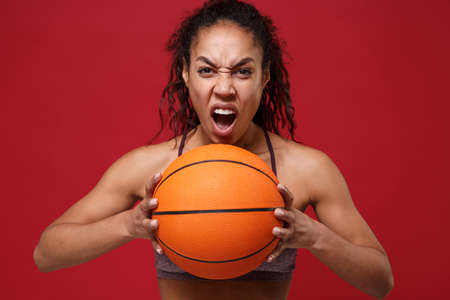Screaming young african american sports fitness basketball player woman in sportswear working out isolated on red background studio portrait. Sport exercises healthy lifestyle concept. Holding ball.の写真素材