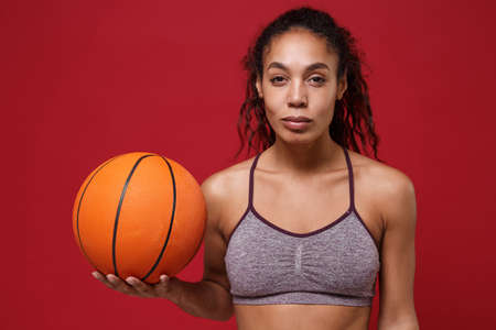 Beautiful young african american sports fitness basketball player woman in sportswear working out isolated on red background studio portrait. Sport exercises healthy lifestyle concept. Holding ball.の写真素材