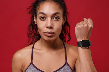 Close up of young african american sports fitness woman in sportswear working out isolated on red background. Sport exercises healthy lifestyle concept. Wearing on hand smart watch with blank screen.の写真素材