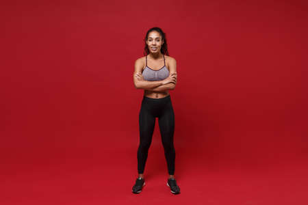 Smiling young african american sports fitness woman in sportswear posing working out isolated on red wall background studio portrait. Sport exercises healthy lifestyle concept. Holding hands crossed.の写真素材