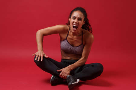 Screaming young african american sports fitness woman in sportswear posing working out isolated on red wall background. Sport exercises healthy lifestyle concept. Sitting, touching knee, get injured.の写真素材