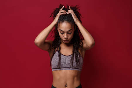 Beautiful young african american sports fitness woman in sportswear posing working out isolated on red wall background studio portrait. Sport exercises healthy lifestyle concept. Put hands on head.の写真素材