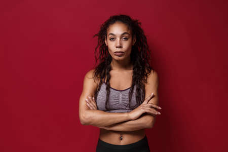 Stunning young african american sports fitness woman in sportswear posing working out isolated on red wall background studio portrait. Sport exercises healthy lifestyle concept. Holding hands crossed.の写真素材
