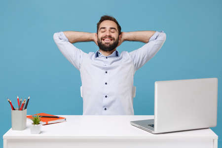 Relaxed young man in light shirt sit work at desk with pc laptop isolated on pastel blue background. Achievement business career lifestyle concept. Keeping eyes closed sleeping with hands behind head.の写真素材