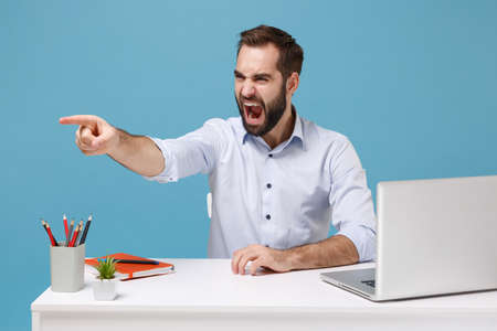 Angry young man in shirt sit work at desk with pc laptop isolated on blue background. Achievement business career lifestyle concept. Mock up copy space. Pointing index finger aside screaming swearing.の写真素材